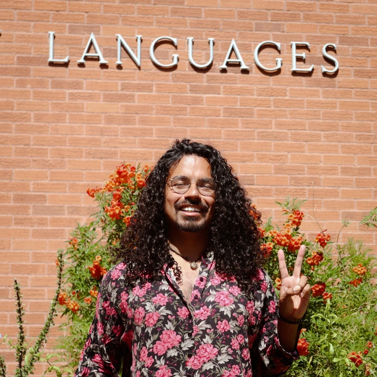 Image of professor, standing in front of brick building, professor is holding the peace sign with their hand